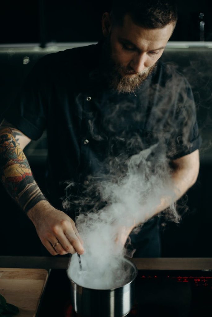 pexels photo 3338535 A bearded chef in a black uniform cooking with steam rising in a modern kitchen.